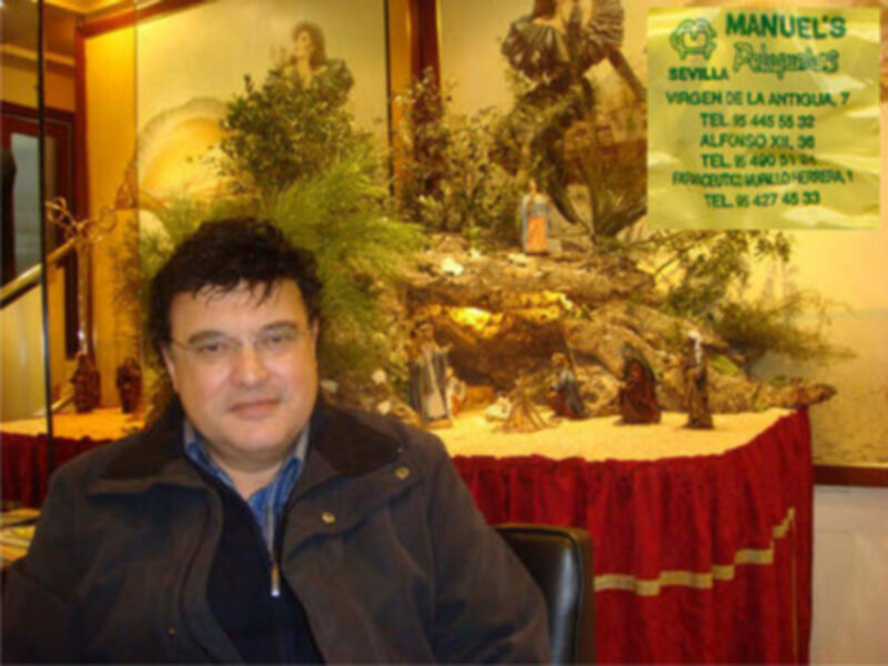 Un hombre con una chaqueta marrón y una camisa azul está sentado frente a un escaparate de una tienda. La imagen muestra una variedad de figuras religiosas y un cartel con información sobre la tienda.