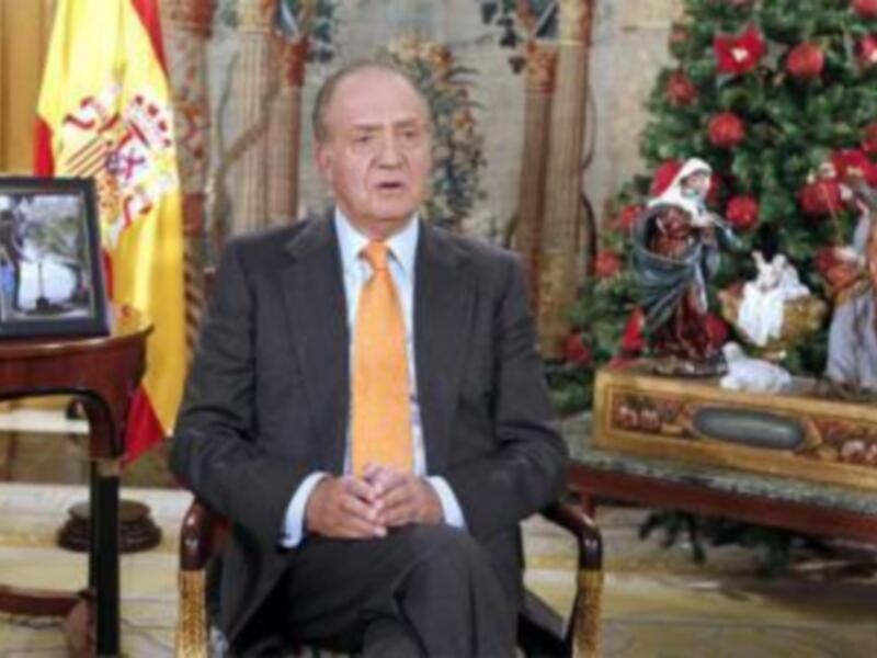Persona en traje y corbata sentada en un sillón, fondo con bandera española y decoración navideña.