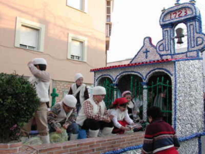 Una escena festiva con personas vestidas de blanco y negro frente a una estructura artística adornada con flores. La imagen se sitúa en un entorno urbano, frente a una casa con ventanas y fachada.