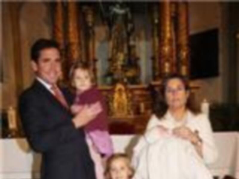 Una familia feliz durante un evento religioso, con una niña en el centro y dos adultos a sus lados. La escena se desarrolla en un ambiente festivo, con una mesa y decoración al fondo.