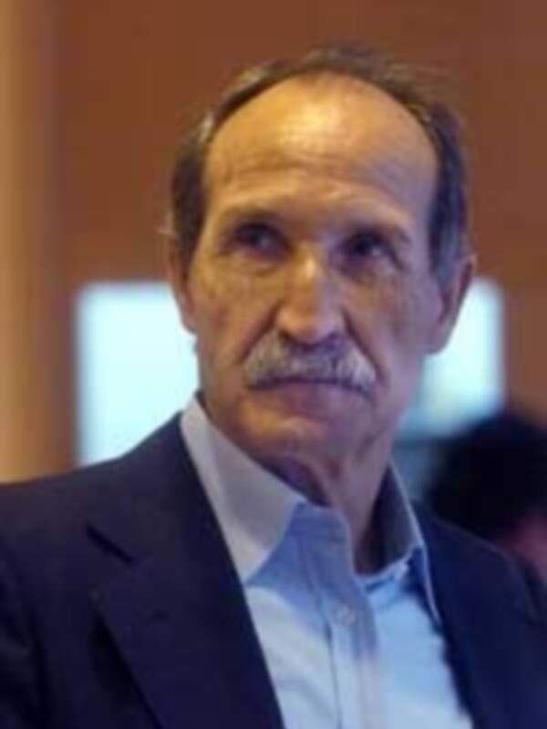 Un hombre con barba y bigote, vestido con una chaqueta negra sobre una camisa azul claro.