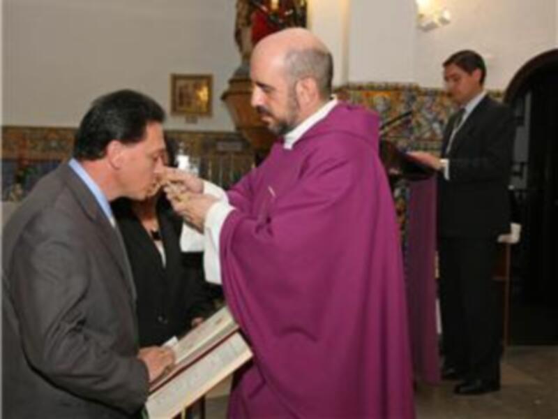 Un sacerdote en un hábito rojo bendice a una persona con un vaso de agua, mientras otro hombre observa. La imagen sugiere una ceremonia religiosa o ritual.
