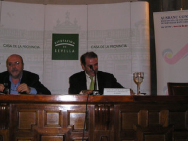 Dos hombres sentados en un escenario, con una pantalla de fondo que muestra el logo de la Diputación de Sevilla y el texto 'Casa de la Provincia'.