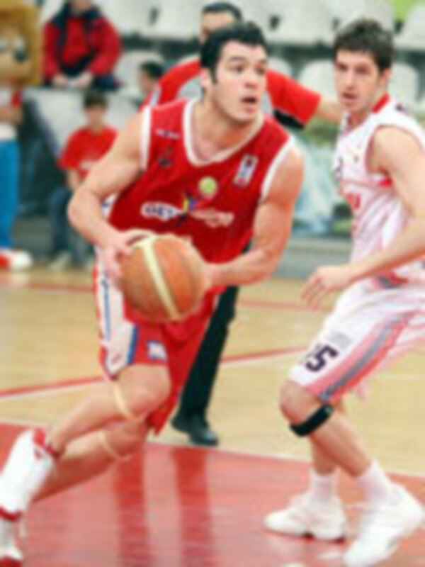 Jugadores de baloncesto en acción durante un partido, con un jugador en rojo y otro en blanco, ambos con el balón y una pelota en el aire.
