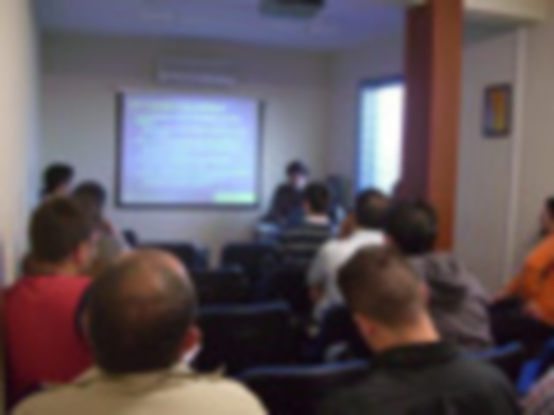 Un grupo de personas en una sala de conferencias, escuchando a un orador frente a una gran pantalla proyectada. La imagen transmite la idea de educación o formación en un entorno profesional.