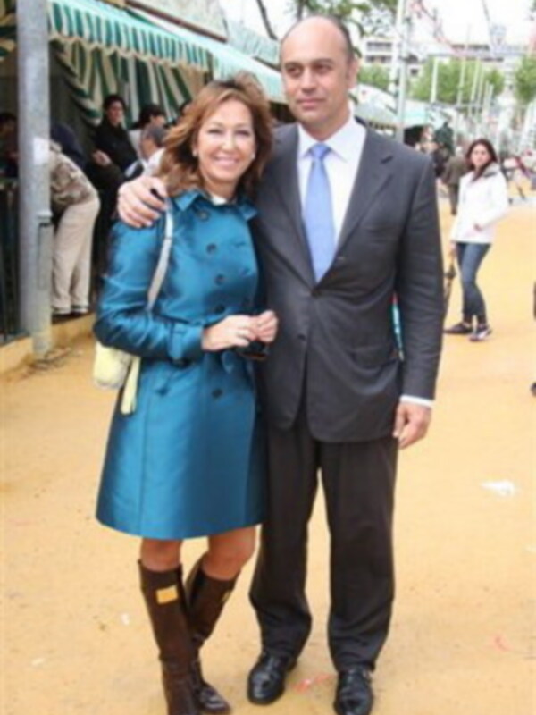 Una pareja posando juntos en un evento al aire libre, con la mujer vestida con una chaqueta azul y botas altas, y el hombre con una chaqueta negra y corbata azul. El fondo muestra un ambiente festivo con toldos y personas en el horizonte.