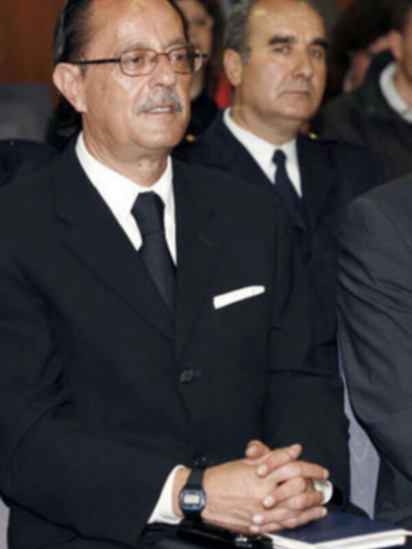 Un hombre en traje oscuro con corbata y reloj de pulsera, junto a otros individuos en una reunión formal.