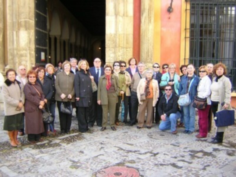 Una multitud de personas en trajes formales y chaquetas posan frente a una entrada monumental, con un hombre en el centro usando gafas de sol y una chaqueta azul. La imagen muestra una escena formal en un entorno histórico con detalles arquitectónicos.