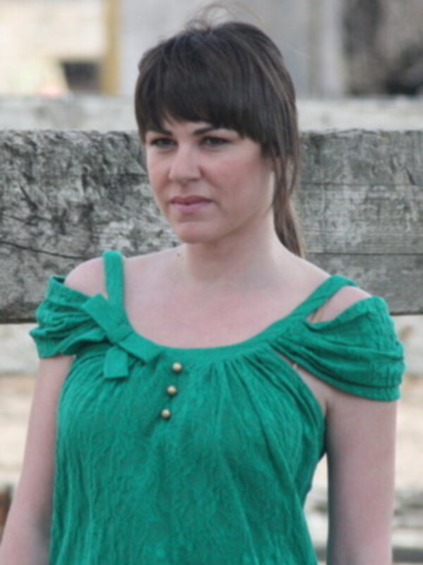 Una mujer con cabello oscuro, mirando directamente a la cámara, vistiendo una camiseta verde con botones dorados y mangas holgadas.