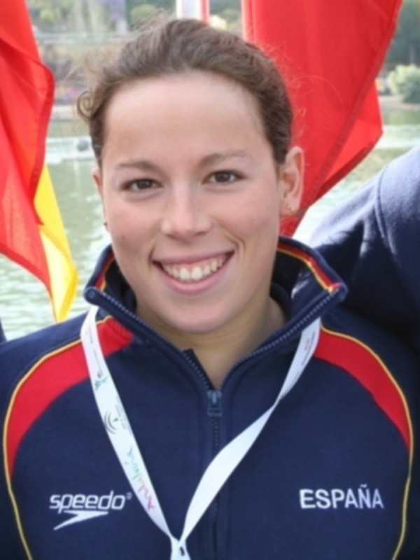 Joven mujer sonriente con medalla y chaqueta de natación española, frente a una bandera española.