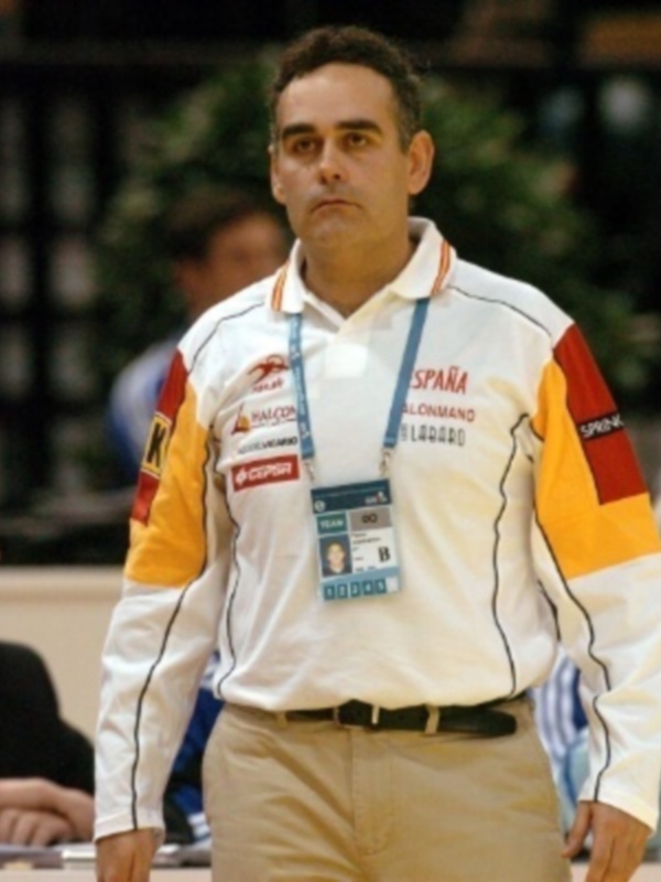 Un hombre con una camisa deportiva de color blanco y amarillo con detalles en rojo, lleva un cinturón negro y una identificación azul. Está en un entorno con otros individuos, posiblemente en una conferencia o evento.