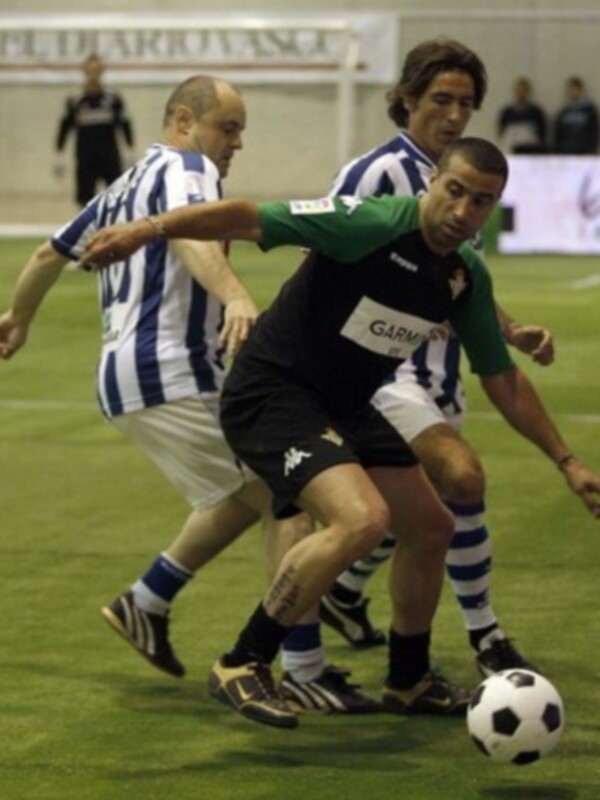 Tres jugadores de fútbol en acción, uno con camiseta verde y otro con blanco y negro, disputan el balón en un campo de fútbol.