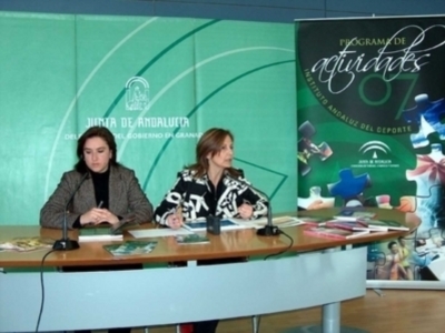 Dos mujeres sentadas en una mesa de conferencia, frente a un fondo verde con el logo del Junta de Andalucía y un cartel que anuncia actividades deportivas.