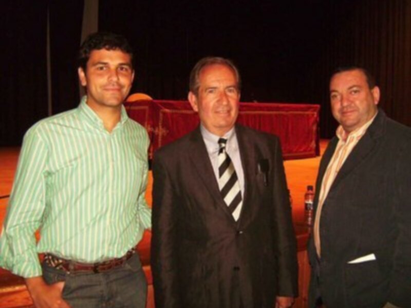 Tres personas posando juntas en un evento formal, con un fondo de cortinas rojas y una persona en el centro con una corbata negra y blanca.