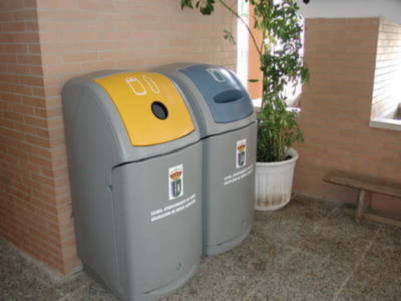 Dos contenedores de reciclaje con un logo y una etiqueta en la pared. Un macetero con una planta y un banco de madera a la derecha.