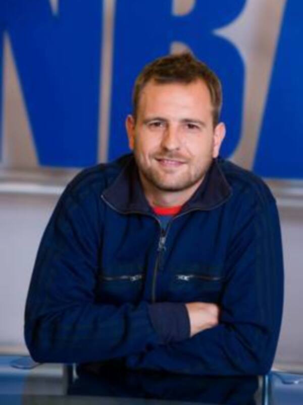 Un hombre con una chaqueta azul de cuello alto y una camiseta roja, con una pared azul en el fondo.