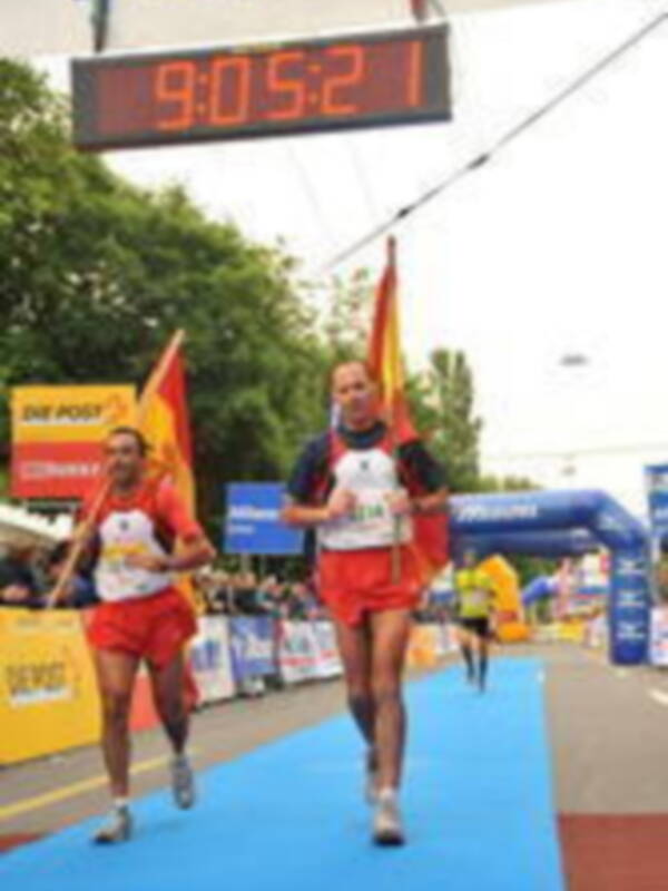 Dos corredores cruzan la línea de meta en una carrera, con un marcador que muestra 9:05:21. La imagen se desarrolla en un entorno verde con árboles y una bandera alemana ondeando.
