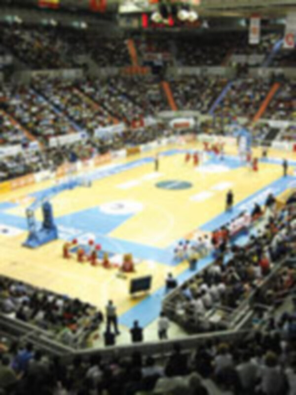 Una imagen de un estadio deportivo lleno con una cancha de baloncesto. Los jugadores están en acción, y la arena está repleta de espectadores. La iluminación es brillante, destacando el centro del campo y los jugadores en movimiento.