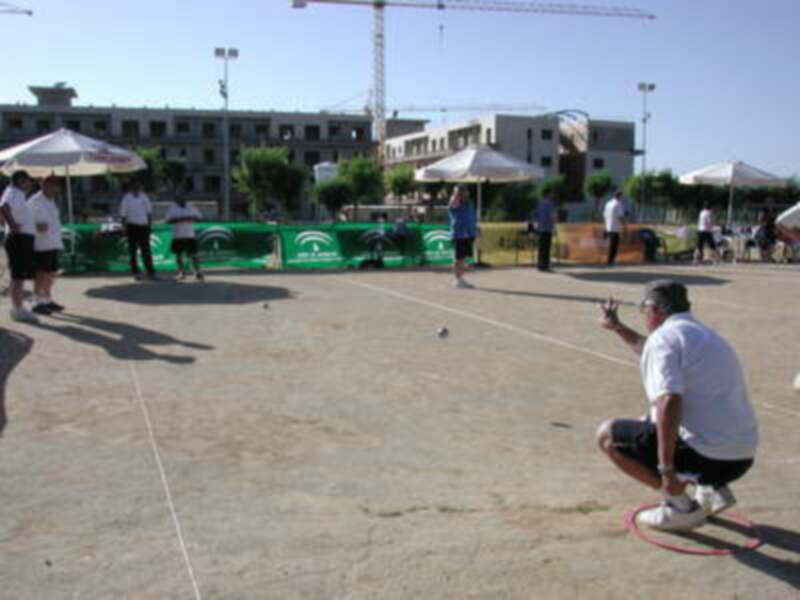 Un hombre en un camiseta blanca y pantalones cortos está lanzando una pelota de ping pong en un campo de tenis. Hay otros jugadores y espectadores alrededor, con sombrillas y un edificio en el fondo.