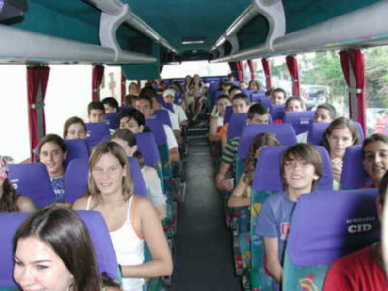 Un grupo de personas sentadas en un autobús lleno, con asientos individuales y una puerta a la izquierda. La imagen muestra un interior de autobús con asientos azules y rojos, algunos pasajeros sonriendo o mirando hacia el frente.