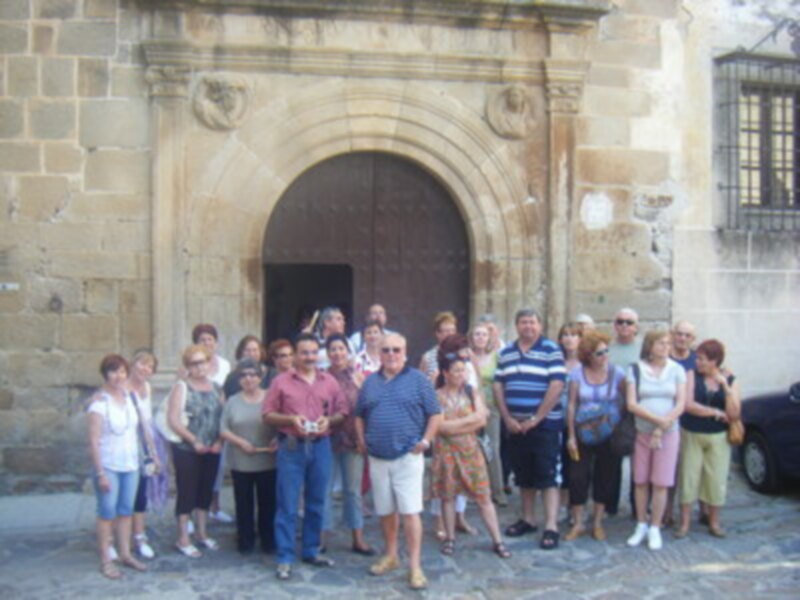 Una imagen de un grupo de personas posando frente a una antigua puerta de piedra con arco. La gente está vestida casualmente, algunas llevan sandalias y pantalones cortos. Al fondo se puede ver una pared de ladrillo y un edificio con ventanas de hierro.