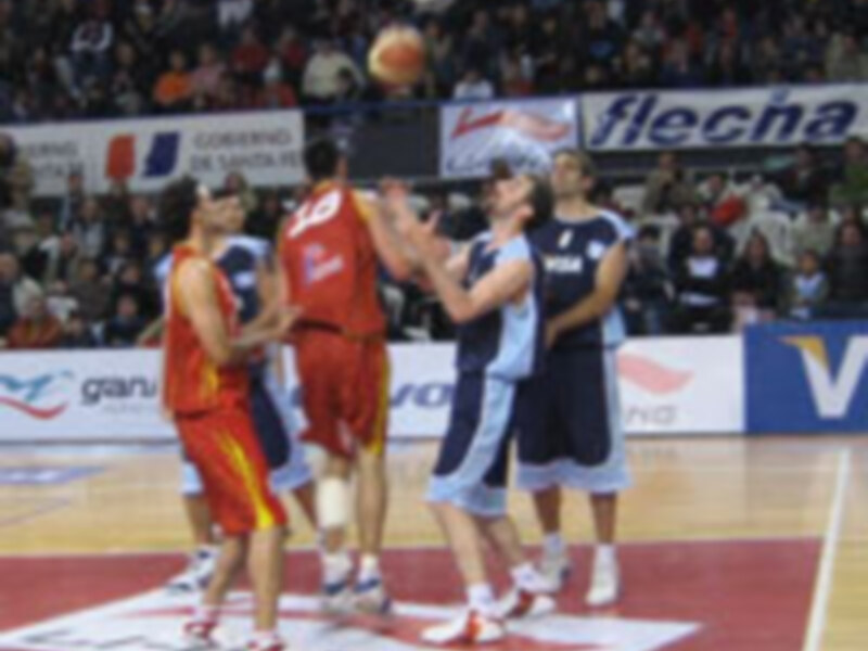 Un juego de baloncesto en una cancha con jugadores en acción, uno de ellos con una camiseta roja y otro con una camiseta azul. La publicidad en el fondo incluye 'Flecha' y 'Pepsi'.