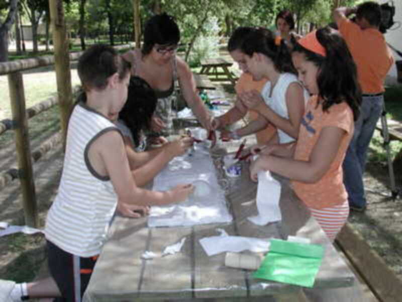 Niños y niñas están sentados en una mesa de picnic, pintando con tizas. La imagen muestra un ambiente familiar y creativo en un parque.