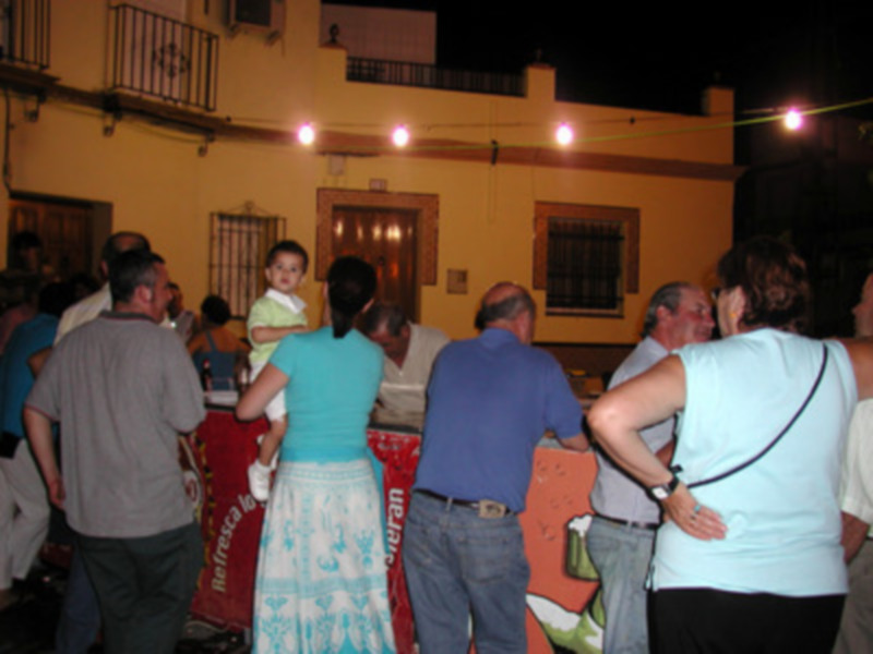Una familia en una fiesta nocturna, con gente de todas las edades disfrutando. La imagen muestra una casa amarilla con luces rojas iluminando la noche, y personas en ropa casual.