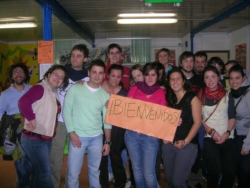 Una multitud de personas sonríe y posa para la foto, con un hombre en el centro sosteniendo una pancarta que dice "¡BIENVENIDOS!". La imagen muestra un ambiente festivo y alegre, con carteles de colores en el fondo.