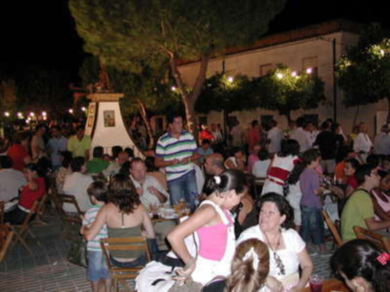 Una noche en un barrio con gente reunida alrededor de mesas, disfrutando de una fiesta. La iluminación es suave y la atmósfera es agradable, con árboles y edificios en el fondo.