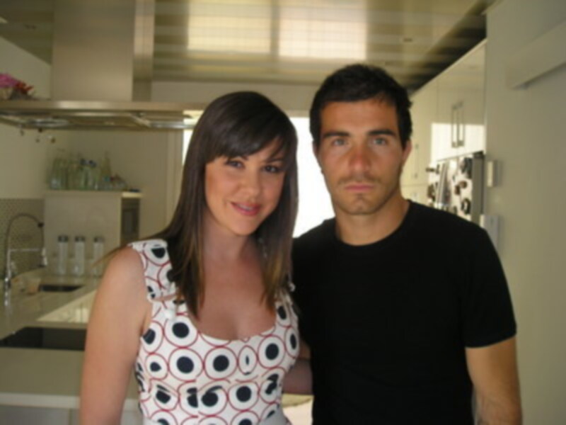 En una cocina moderna, una mujer con cabello largo y una camiseta estampada de círculos blancos y rojos posa junto a un hombre con una camiseta negra. Al fondo, se pueden ver elementos de cocina como frigoríficos y un fregadero.