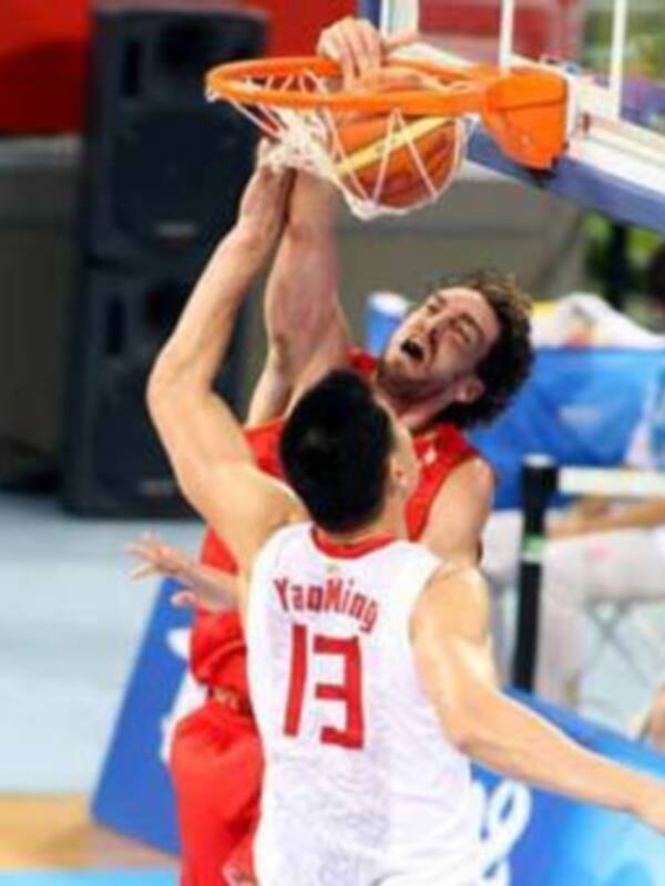 Un jugador de baloncesto chino intenta anotar un tiro libre mientras otro jugador rival lo bloquea. El fondo muestra una cancha de baloncesto con un público en el borde del campo.