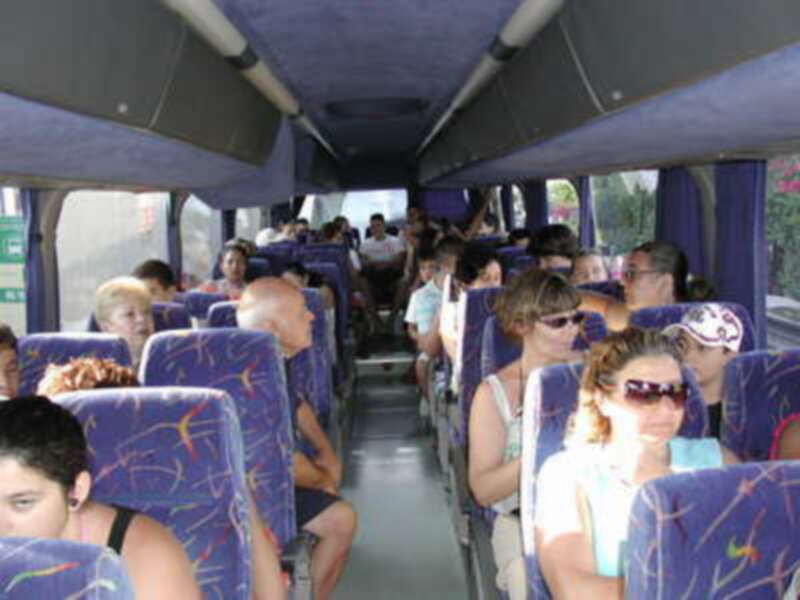 Un autobús lleno de pasajeros sentados en asientos azules con dibujos, mirando hacia adelante. La imagen muestra una variedad de personas, algunas con sombrillas y otros con gafas. La iluminación es natural, sugiriendo que es un día soleado.