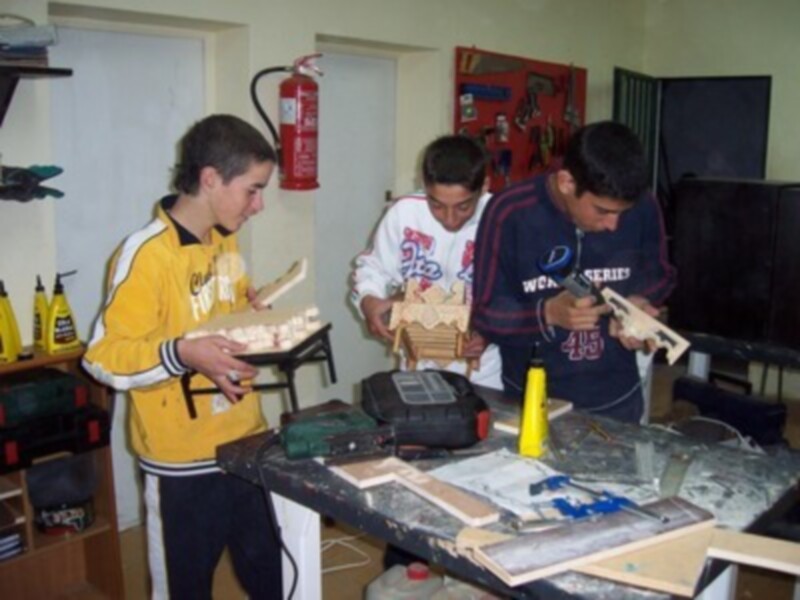 Tres jóvenes trabajando en un taller, uno con una herramienta de madera, otro con un cuchillo y el tercero con un pañuelo amarillo.
