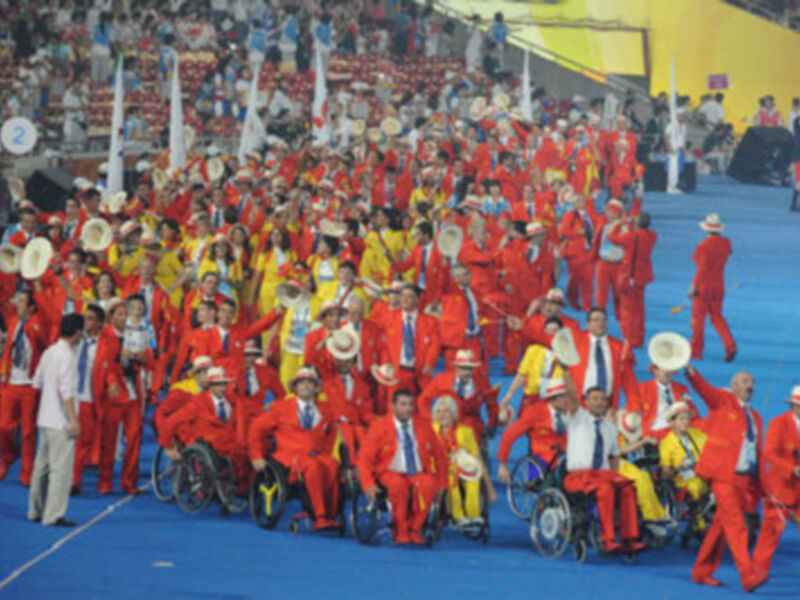Una gran ceremonia de apertura en un estadio lleno, con una multitud de personas vestidas en trajes rojos y amarillos, algunos en sillas de ruedas.
