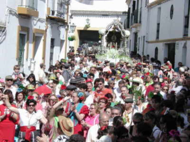 Una procesión religiosa en una calle estrecha, con una imagen de la Virgen en el centro y una multitud de personas vestidas de colores, algunas con sombreros.