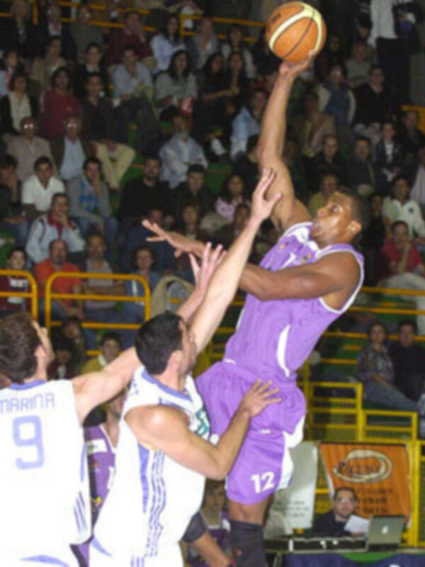 Un jugador de baloncesto en púrpura intenta un tiro libre mientras otros jugadores lo defienden. La arena está llena de espectadores y hay una publicidad en el fondo.