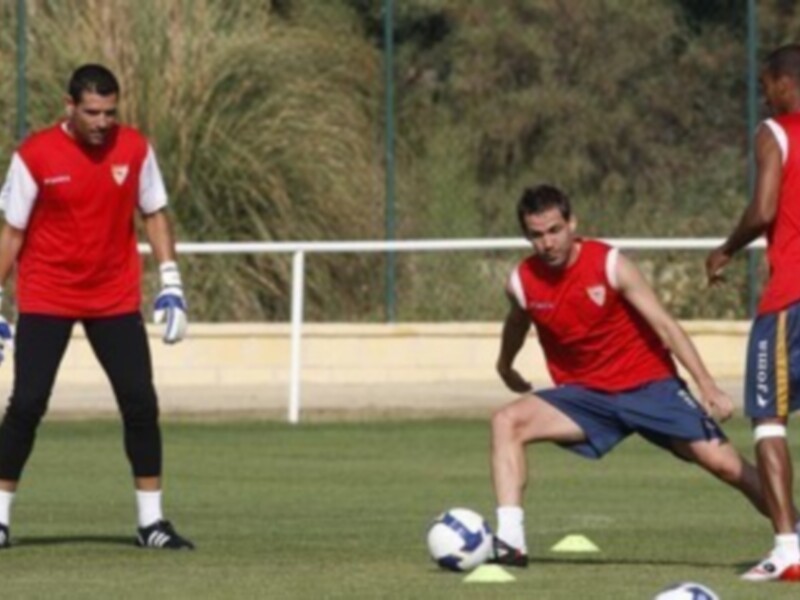Tres jugadores de fútbol realizan ejercicios durante un entrenamiento. El entorno es verde y natural, con una cerca de metal al fondo.