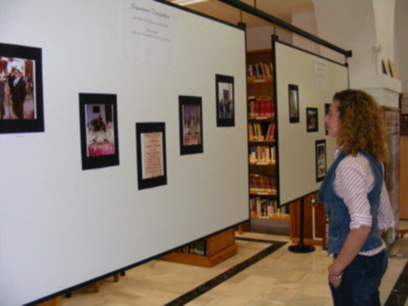Una persona observa una exposición de fotografías en un espacio de exhibición. La pared está llena de fotos y carteles, con una librería en el fondo. La persona lleva un vestido azul y tiene una camiseta blanca por encima.