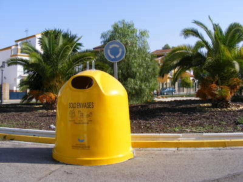 Un contenedor amarillo para residuos orgánicos en un entorno urbano con árboles y edificios al fondo.