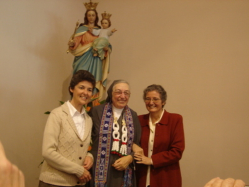 Tres personas sonrientes posan junto a una estatua de la Virgen María con el Niño Jesús. La imagen parece ser de un evento religioso o cultural.