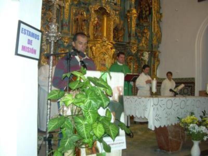 En una iglesia, un hombre habla en un micrófono frente a una imagen religiosa dorada. Detrás de él, otros dos hombres escuchan atentamente. A la derecha, hay flores y una mesa con un cartel informativo.