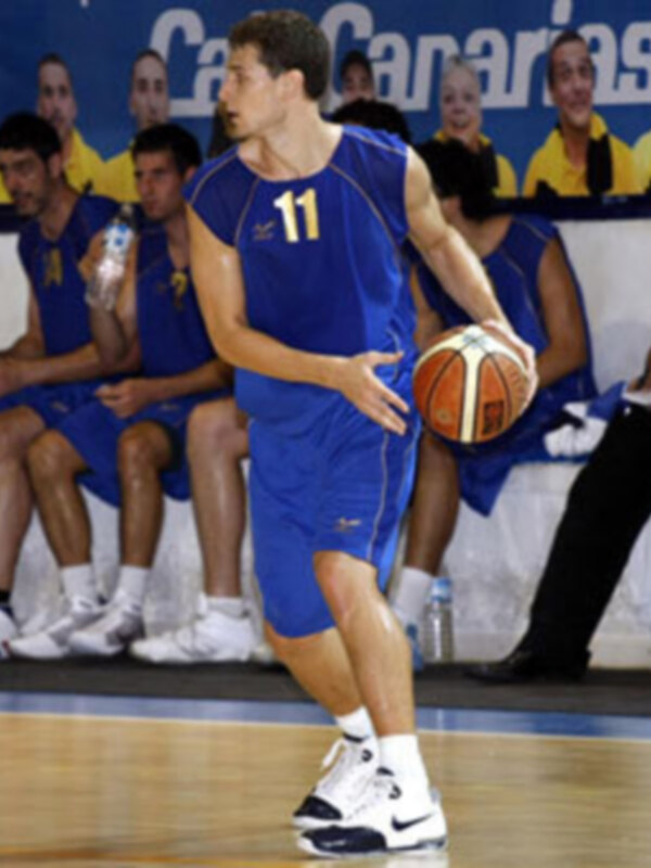 Un jugador de baloncesto en acción, con una camiseta azul y un balón en la mano, mientras un grupo de personas observa desde la banca.