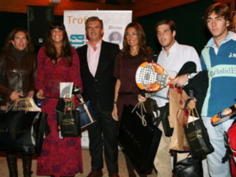 Una imagen de una celebración o evento, con personas posando para la foto. En el fondo hay un cartel que dice "TROFEO". La gente está vestida formalmente, con chaquetas y vestidos. Algunos sujetan bolsas o mochilas, y uno de ellos sostiene un balón. La escena parece ser en una sala o evento al aire libre, con luces y un público.