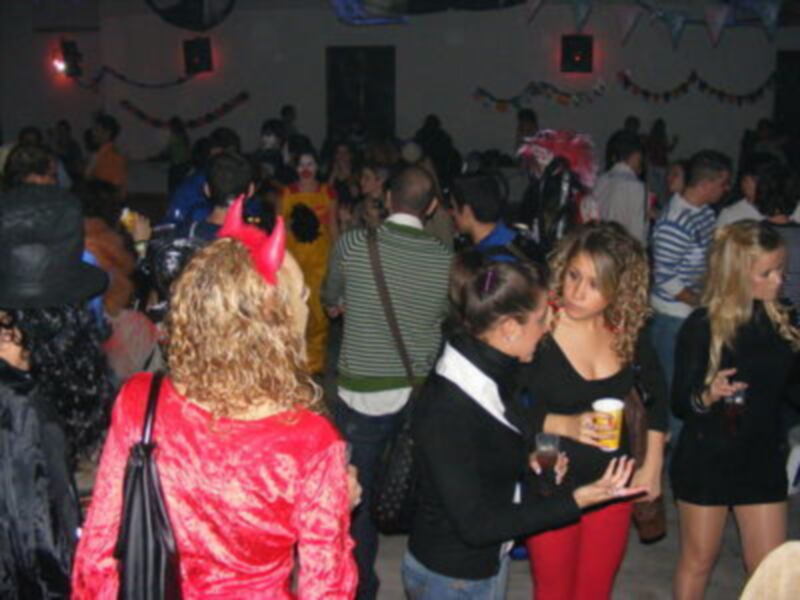 Una fiesta en un salón con gente bailando y bebiendo. La decoración incluye globos y pancartas colgantes.