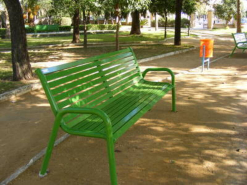 Banco verde en un parque con sombra de árboles, cerca de una mesa y un buzón.