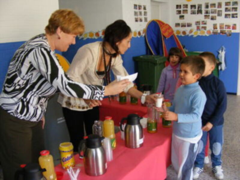 Una persona entrega un documento a un niño mientras otras personas observan. En la mesa hay botellas de leche y jugos. El ambiente parece ser una escuela o centro comunitario, con decoraciones infantiles en el fondo.