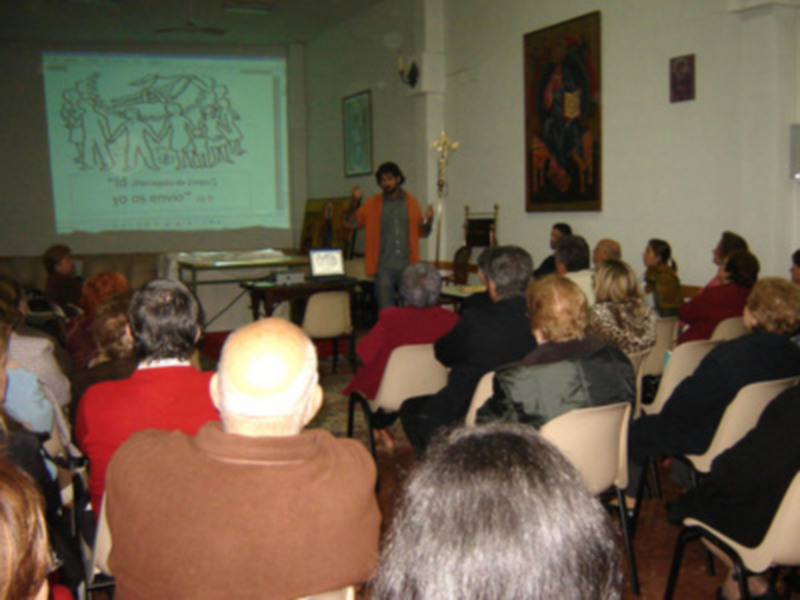 Una sala llena de personas sentadas, viendo una presentación en un proyector. La imagen muestra a una persona hablando frente a la audiencia, con una cruz en el fondo y un cuadro de arte.