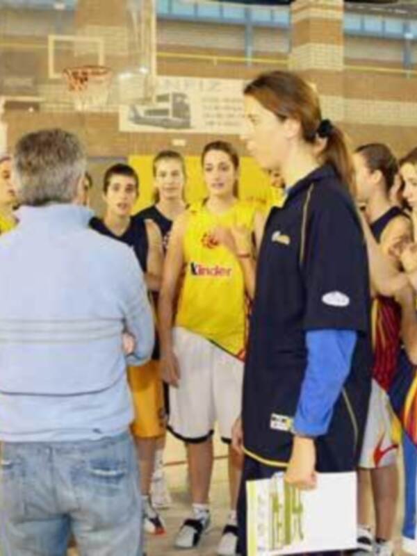 Equipo femenino de baloncesto en una práctica, con una entrenadora y jugadores en el fondo.