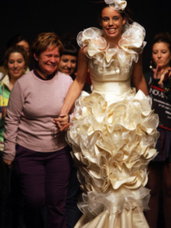 Una modelo posa con un vestido de novia blanco con volantes y una flor en el cabello. La imagen muestra a varias personas observando la presentación de moda.